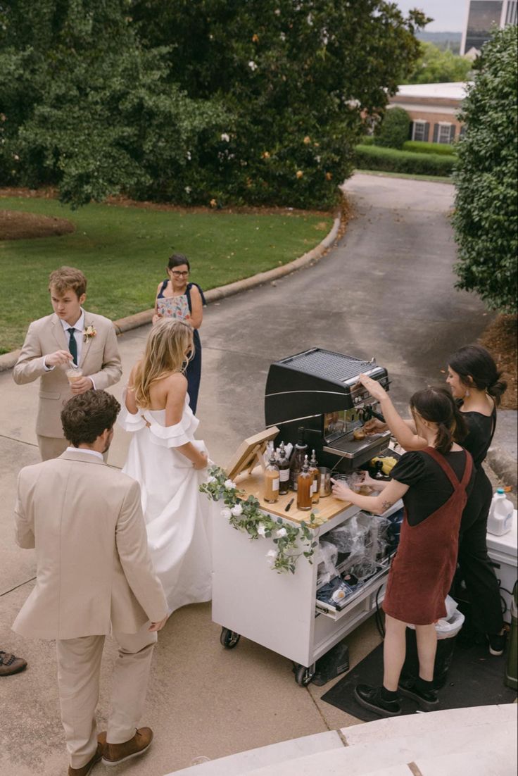 a group of people standing around a table filled with food and drinks on top of a sidewalk