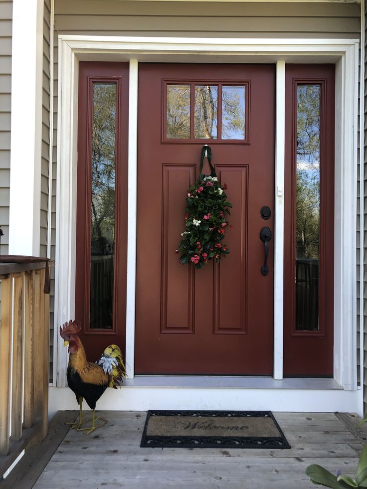 a rooster standing in front of a red door with a wreath on it's side