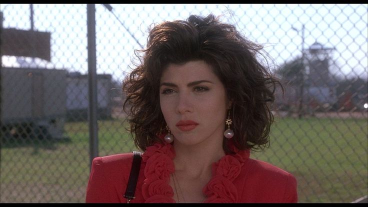 a woman in a red shirt is standing by a fence and looking at the camera