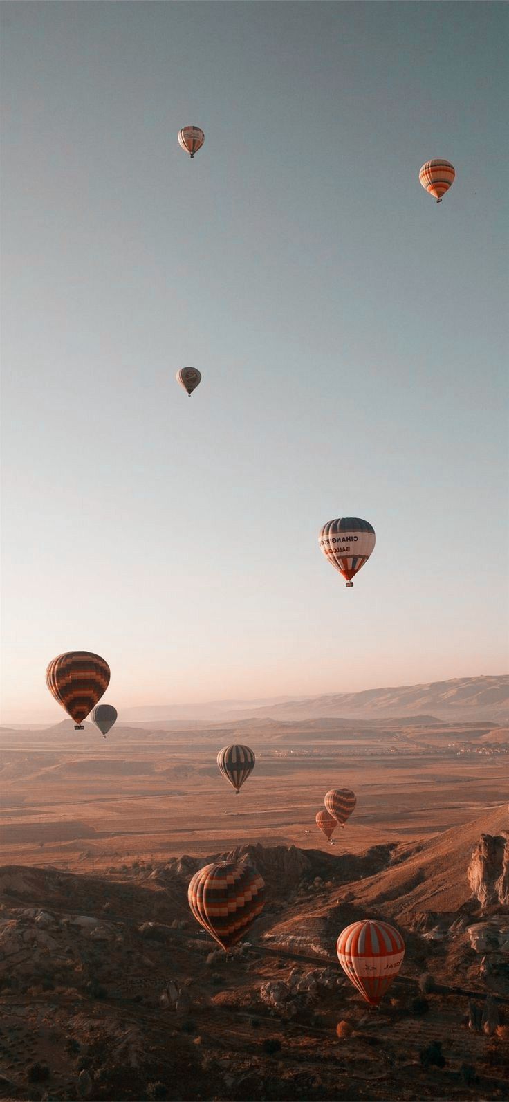 many hot air balloons flying in the sky above some rocks and dirt land with mountains in the background