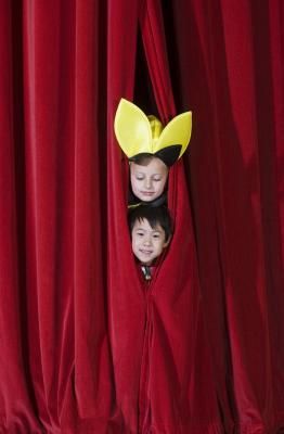 two children are peeking out from behind red curtains with yellow hats on their heads and one child is looking through the curtain