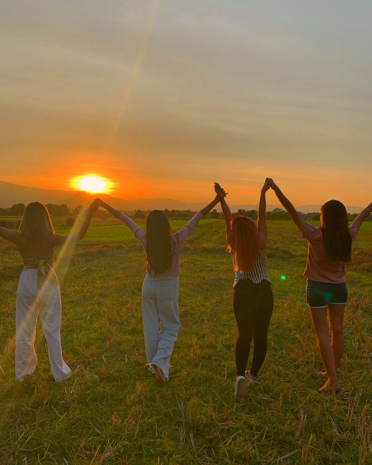 four girls are standing in the grass with their arms up as the sun goes down