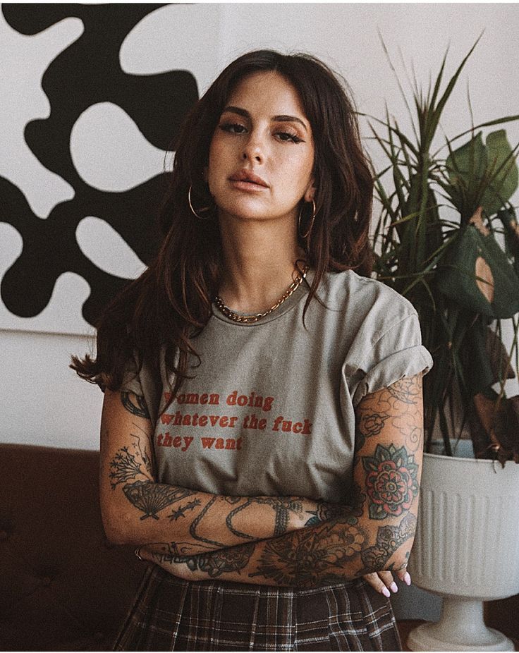 a woman with tattoos standing in front of a potted plant and looking at the camera