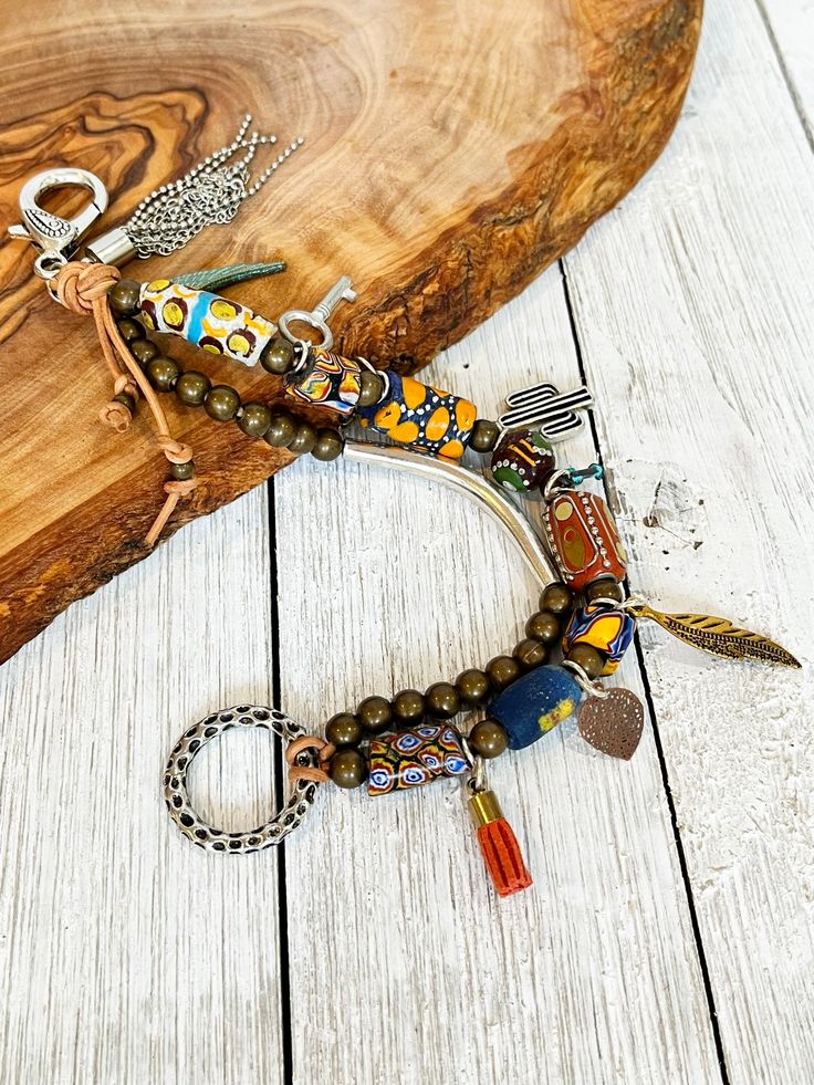 A beautiful boho beaded bracelet featuring gorgeous African trade beads. The trade beads are strung on tan leather cord along with brass beads and assorted charms including a wing, key, elephant, cactus, leaf and small leather tassel. Another strand features brass beads and a curved silver plated tube bead also strung on tan leather cord. Fastens with a silver plated lobster clasp with a silver tassel, and hammered silver ring. The bracelet measures about 8.5 inches around the wrist. African Beaded Jewelry, African Trade Bead Jewelry, Leather Cord Jewelry, Beaded Braclets, Horn Earrings, Hammered Silver Ring, Cord Jewelry, African Trade Beads, Brass Beads