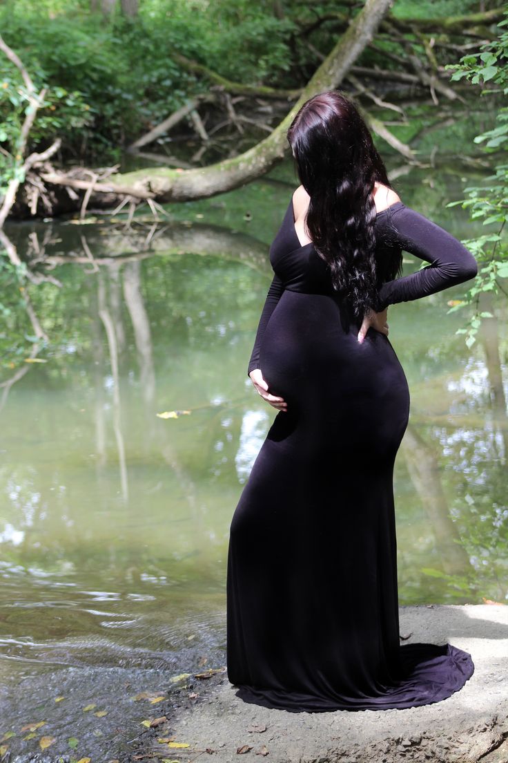 a pregnant woman in a long black dress standing on the edge of a body of water