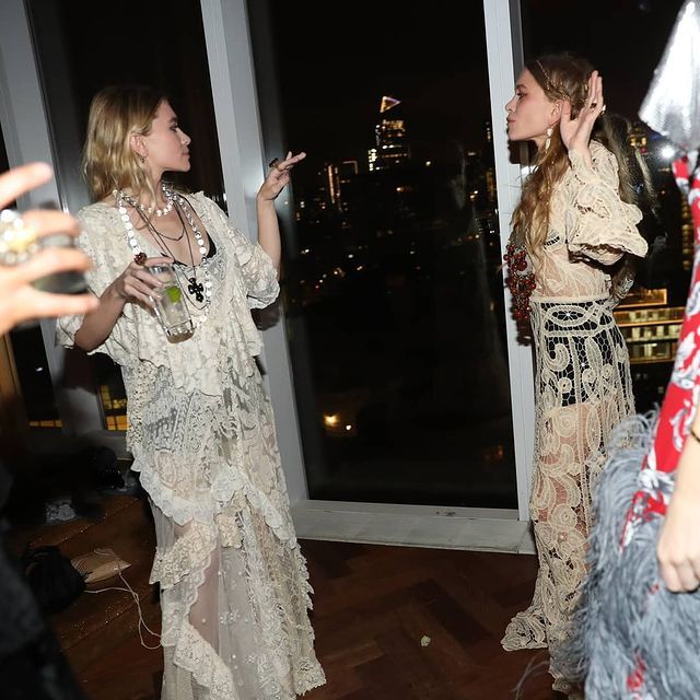 two women standing next to each other in front of a window at night with the city lights behind them