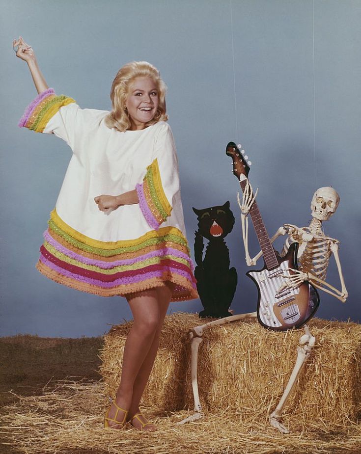 a woman standing next to a skeleton with a guitar and cat on hay bales