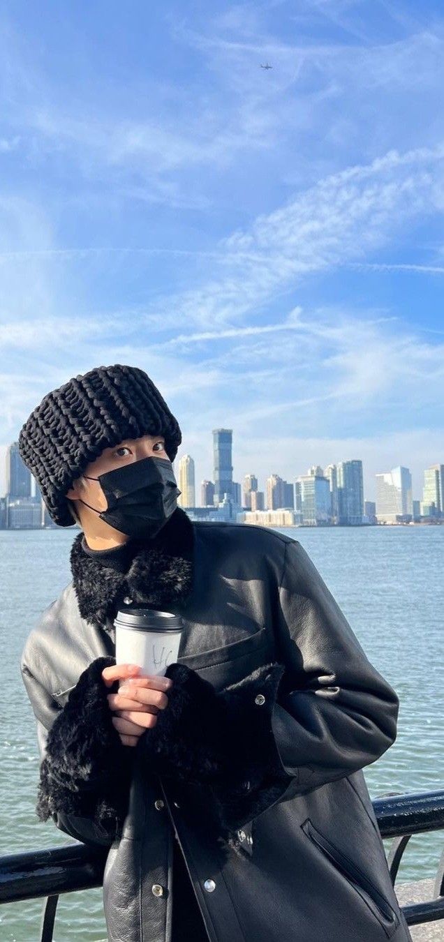 a woman wearing a mask and holding a cup while standing next to the water with a city skyline in the background