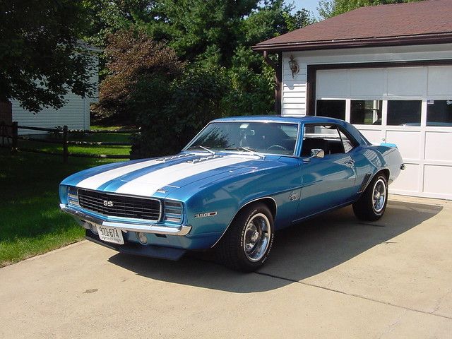 a blue and white muscle car parked in front of a garage with two doors open