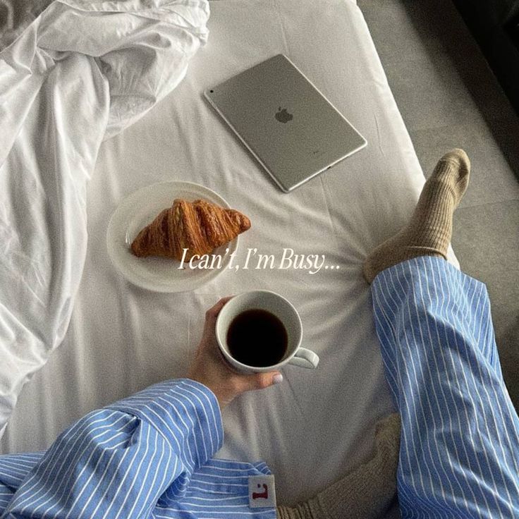 a person sitting on a bed holding a cup of coffee and an apple macbook