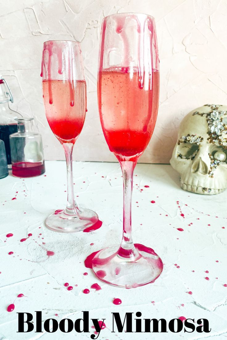 two wine glasses filled with pink liquid sitting on top of a table next to a skull