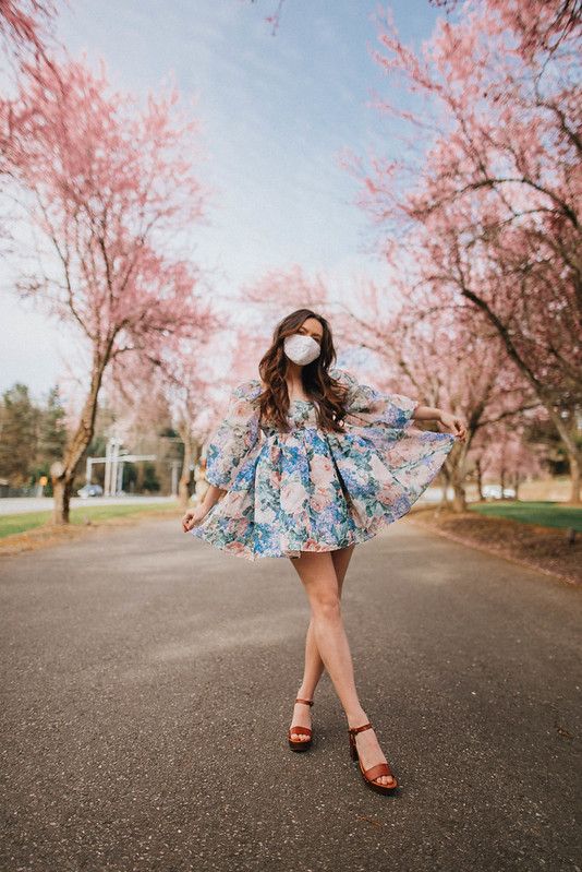 a woman wearing a mask standing in the middle of a road with cherry blossom trees behind her