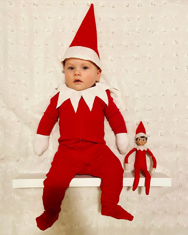 a baby wearing a red and white elf outfit sitting on a shelf next to a stuffed toy