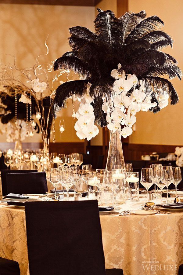 a tall centerpiece with white flowers and feathers sits atop a table set for an elegant dinner