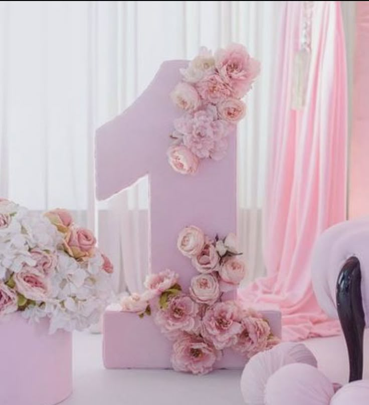 a pink number one surrounded by flowers and balls on a white table with curtains in the background