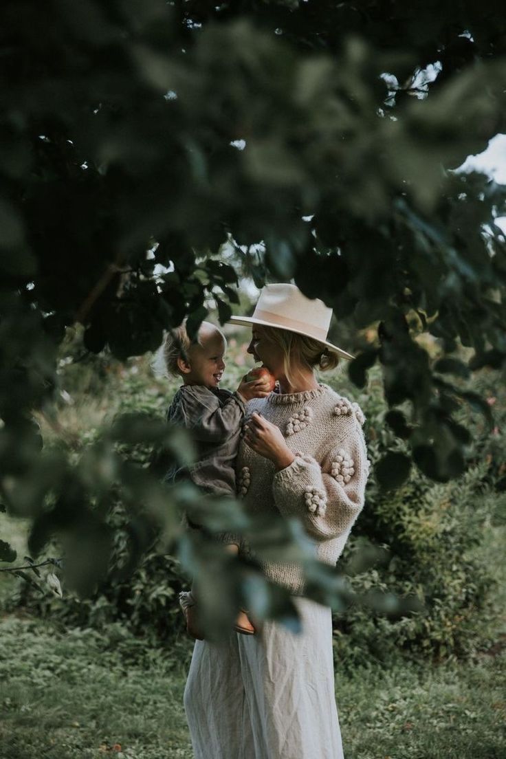 a woman holding a baby in her arms while wearing a white hat and dress standing next to trees