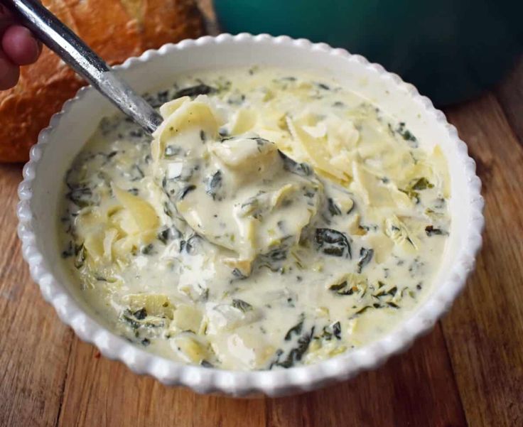 a bowl filled with cheese and spinach on top of a wooden table next to a loaf of bread