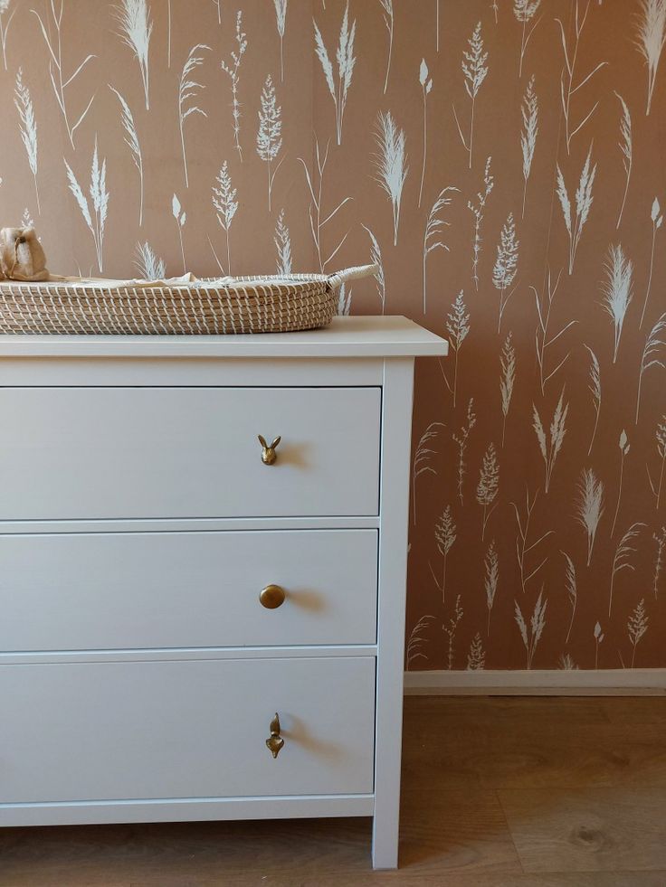 a white dresser with a basket on top of it next to a wallpapered wall