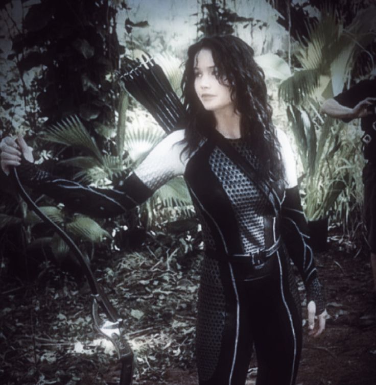 a woman dressed in black standing next to a forest filled with trees and plants, holding her arms out