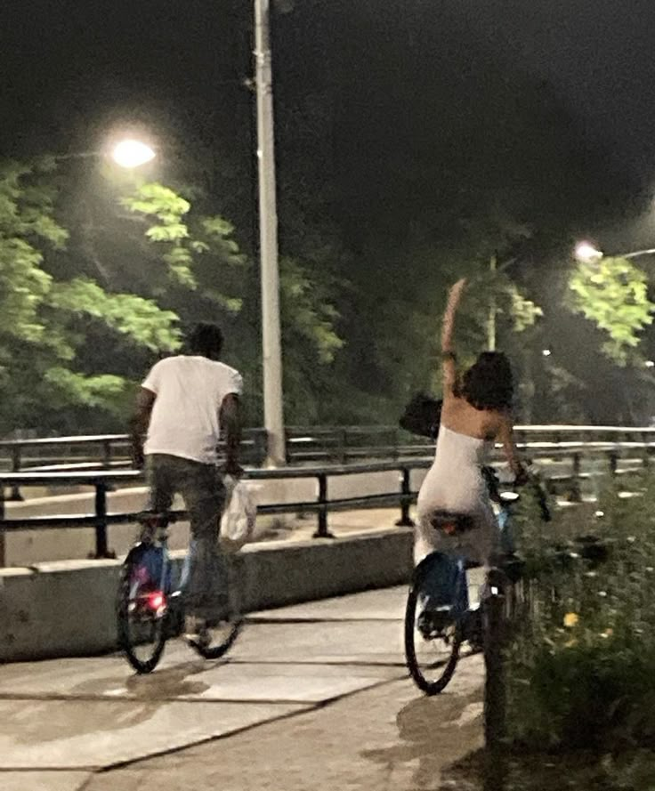 two people riding bikes down a street at night time, one on the back of the bike