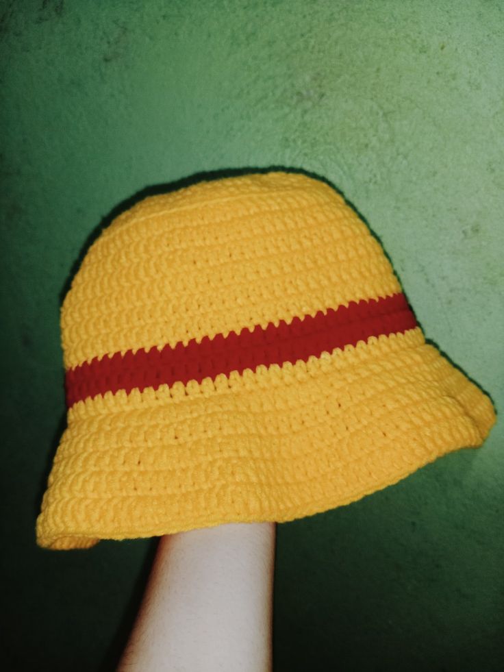 a yellow crocheted hat with red stripes on the brim is held up against a green wall