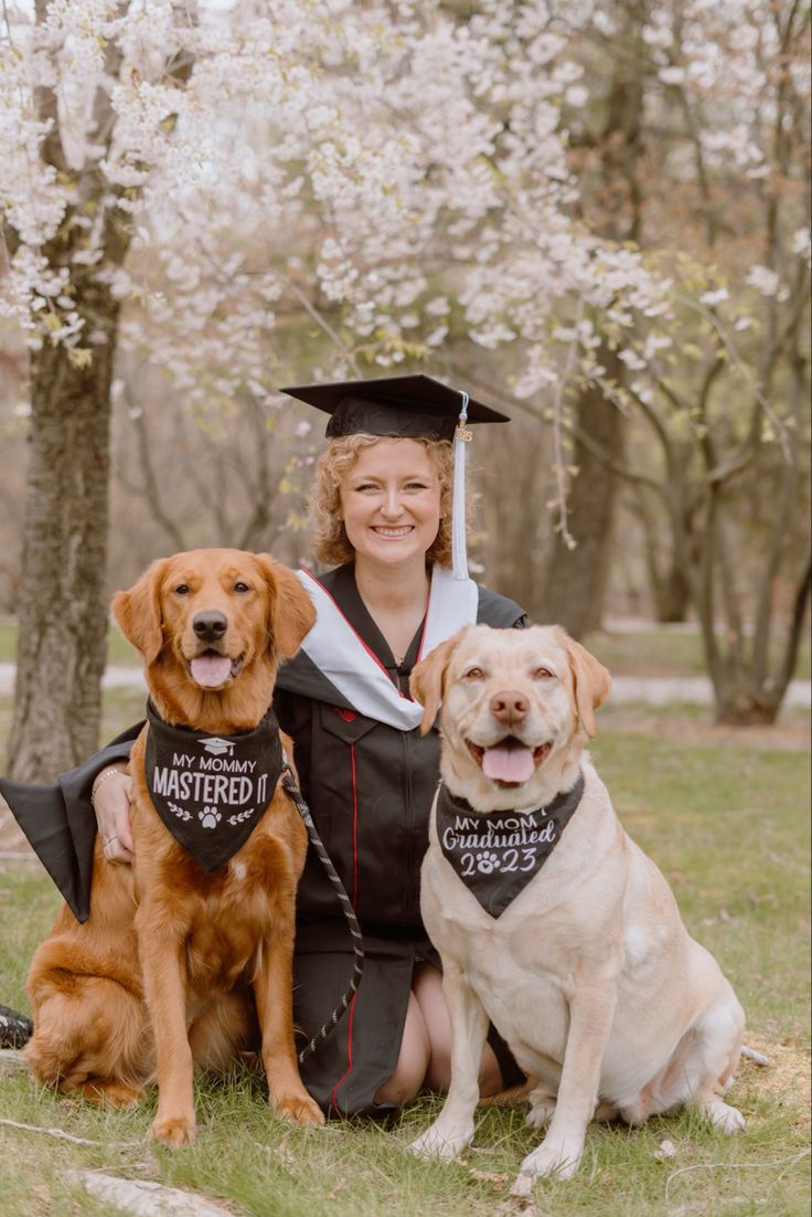 a woman sitting with two dogs in front of cherry trees wearing graduation caps and gowns