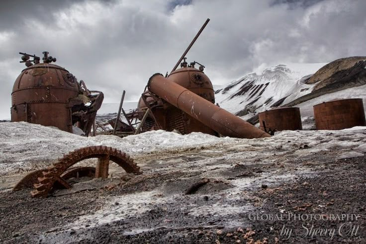 an old rusted out machine in the middle of some snow covered ground with mountains in the background