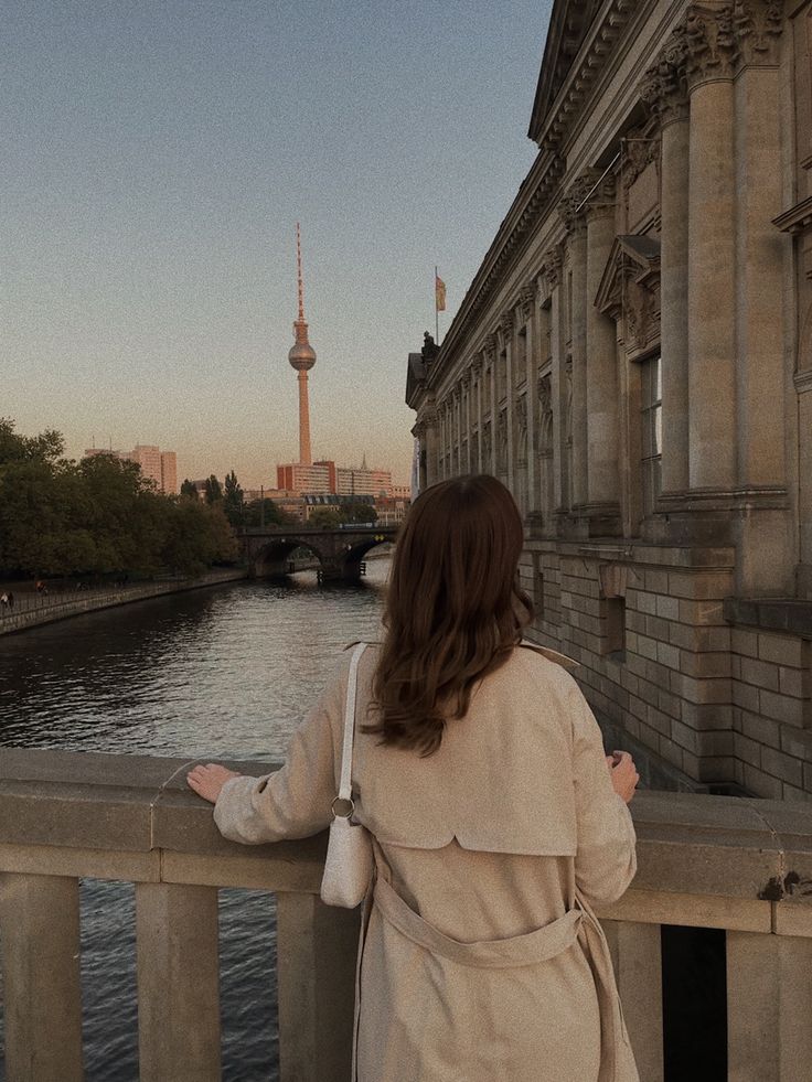 a woman is standing on a bridge looking at the water and buildings in the distance