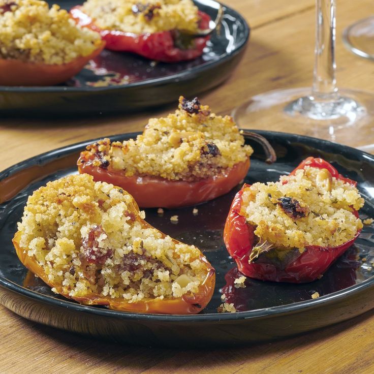 two black plates filled with stuffed peppers on top of a wooden table next to wine glasses