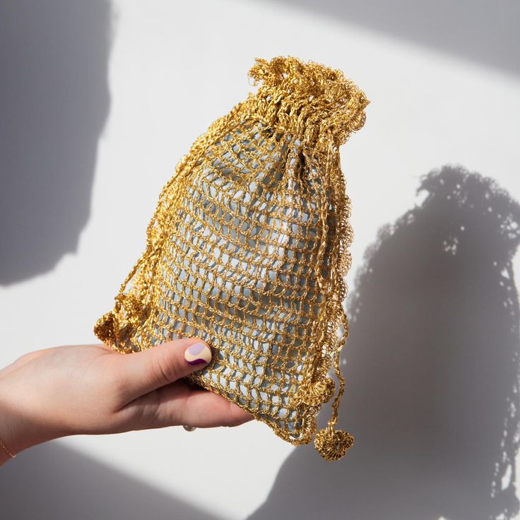 a hand holding a gold colored mesh bag in front of a white wall with shadows on it