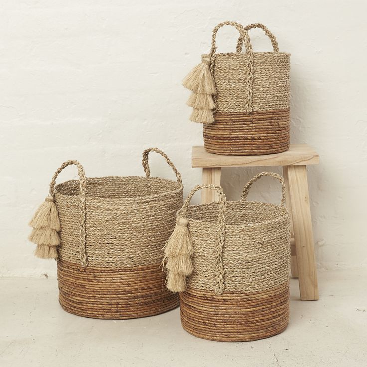 three woven baskets with tassels on them sitting next to a wooden stool against a white wall