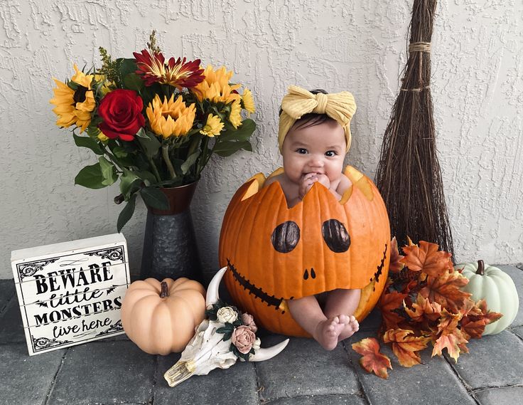 a baby in a pumpkin costume sitting next to some flowers and other halloween decorations on the ground