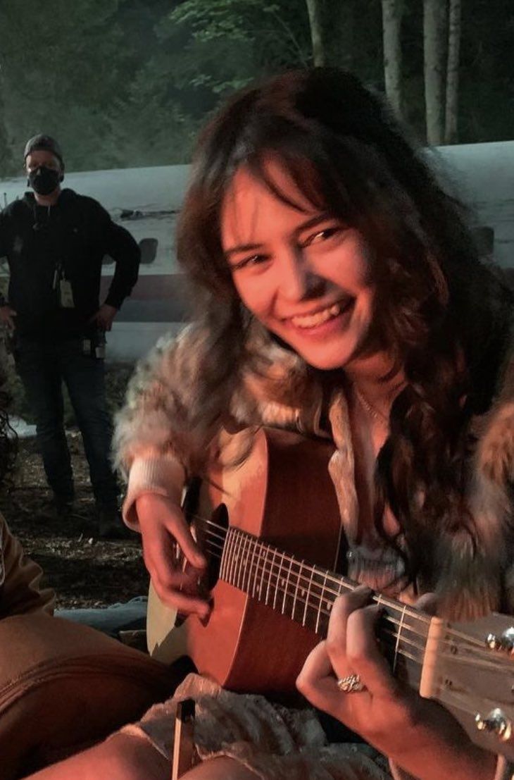 a woman is playing an acoustic guitar while another man stands behind her in the background