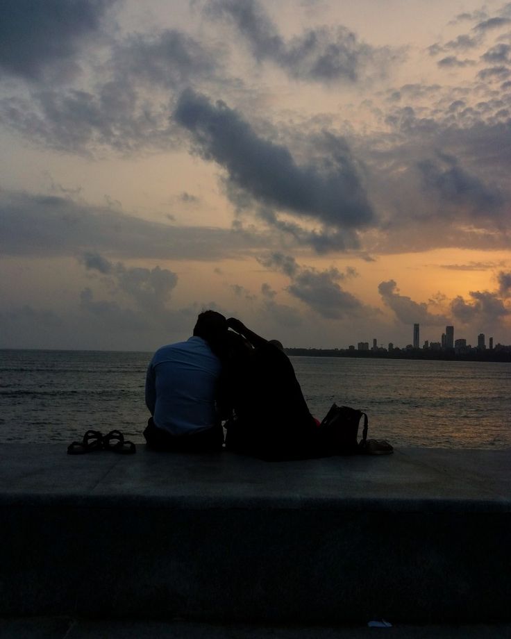 two people sitting on the edge of a pier at sunset with city in the background