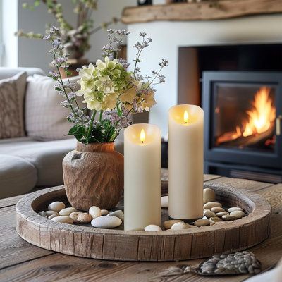 two white candles sitting on top of a wooden tray next to a fire place in front of a couch