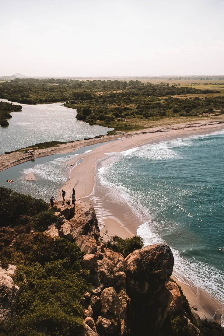 two people walking along the beach next to some water and rocks with trees in the background