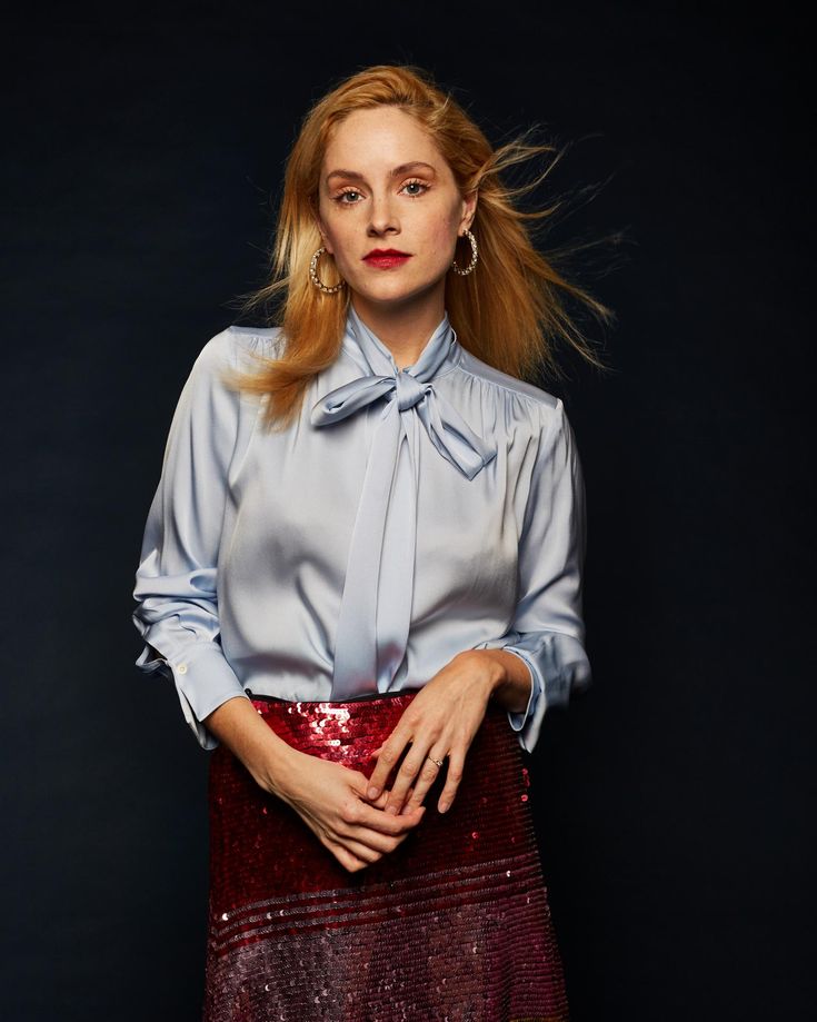 a woman with red hair wearing a blue shirt and maroon skirt standing in front of a black background