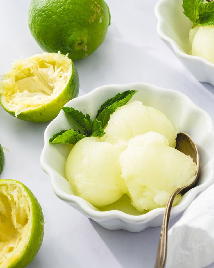 two bowls filled with ice cream next to limes on a white table cloth and spoon