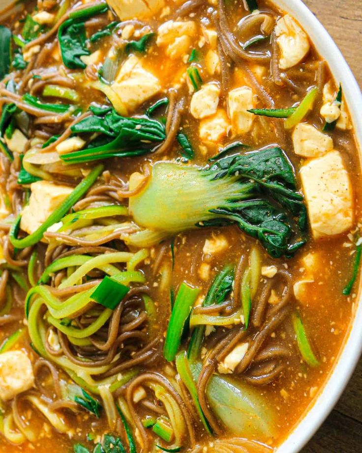 a white bowl filled with noodles, tofu and spinach on top of a wooden table