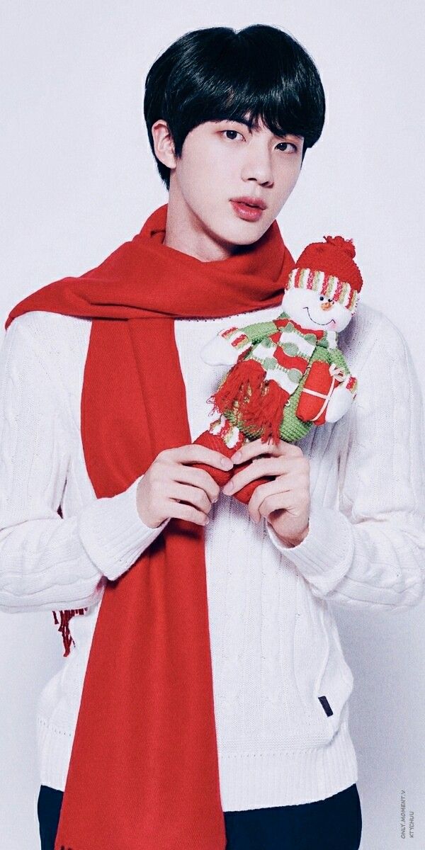 a young man is holding a teddy bear wearing a red scarf and white sweater, while standing in front of a white wall