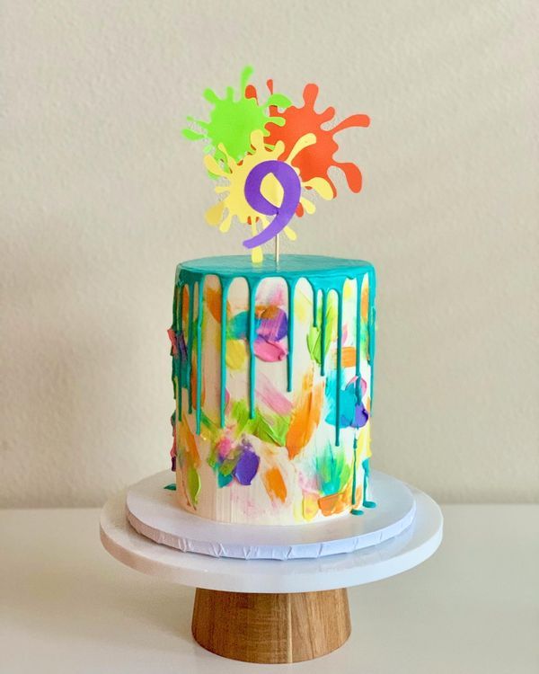 a birthday cake with colorful icing and splattered paint on the top, sitting on a wooden stand