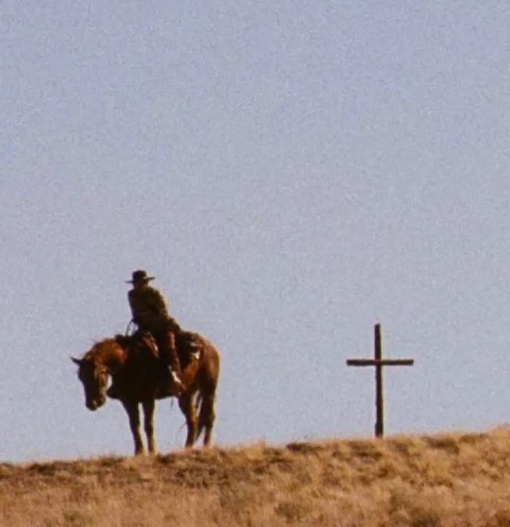 a man riding on the back of a brown horse next to a cross in a field