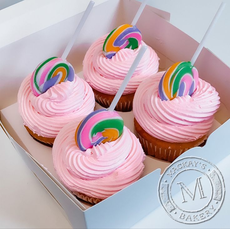 four cupcakes with pink frosting and rainbow icing on sticks in a box