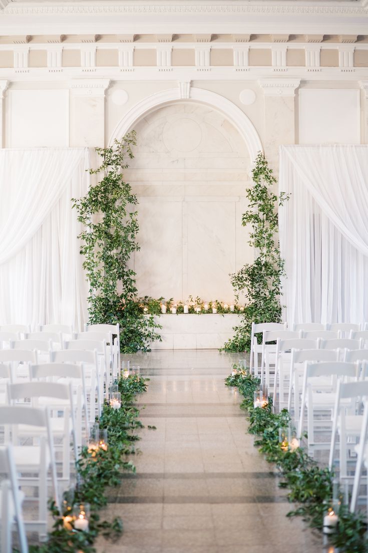 an indoor wedding ceremony with white chairs and greenery on the aisle, decorated with candles