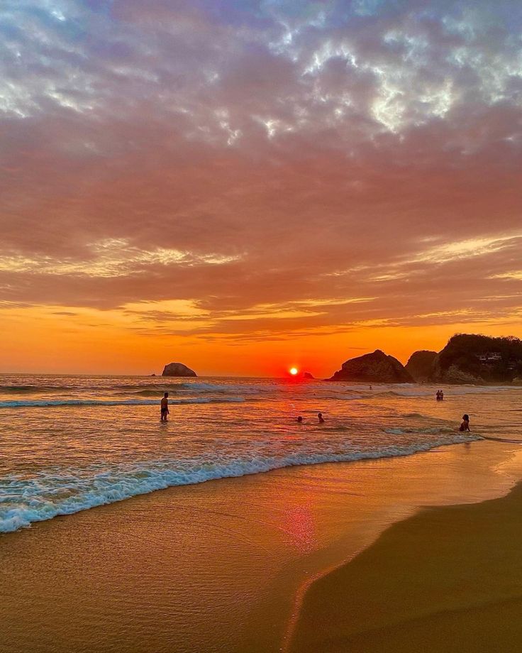 the sun is setting at the beach with people in the water and on the sand