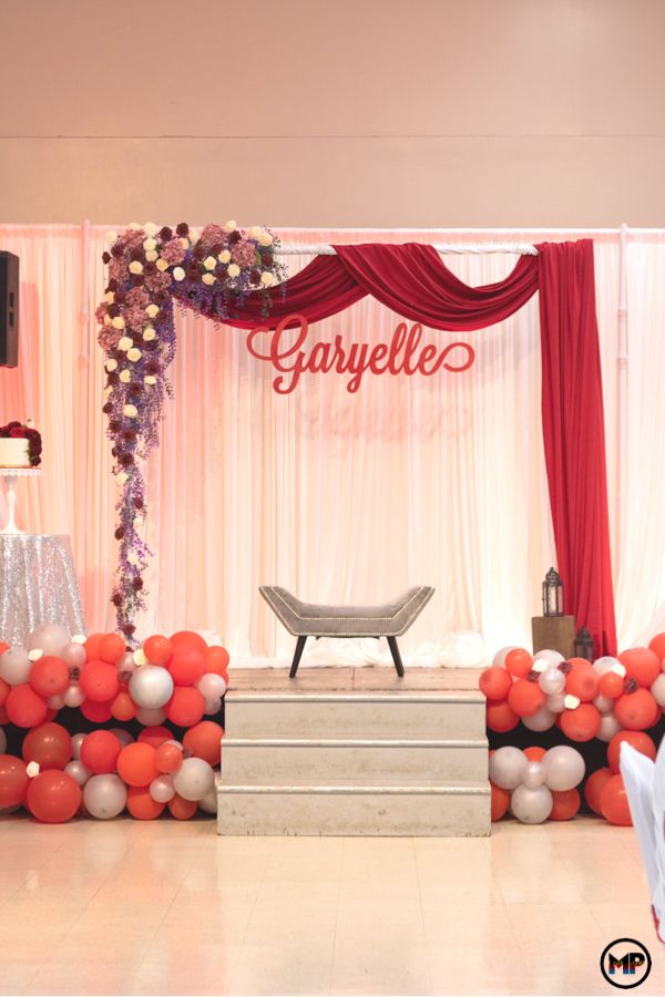 a stage set up for a wedding with red and white balloons on the floor, draped drapes, and flowers