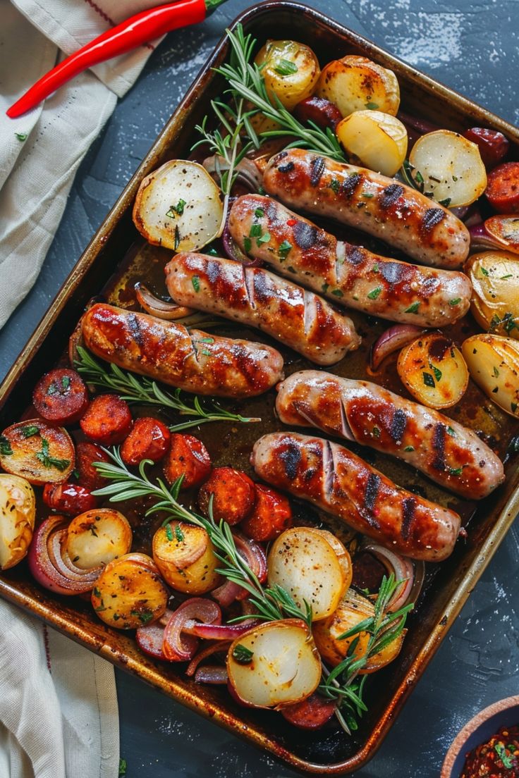 sausages, potatoes and tomatoes in a baking pan on a blue surface with a red pepper