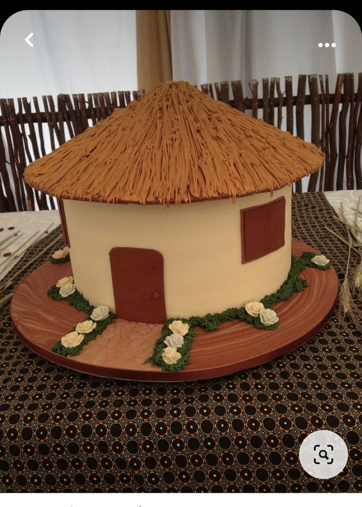 a cake shaped like a house on a plate with flowers around the base and thatched roof