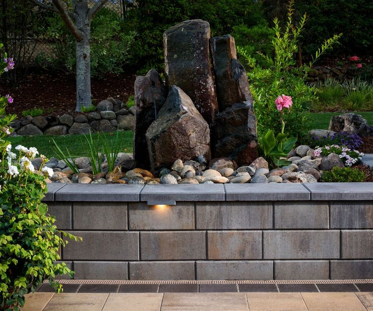 a stone wall with rocks and flowers in the garden next to it is lit up at night