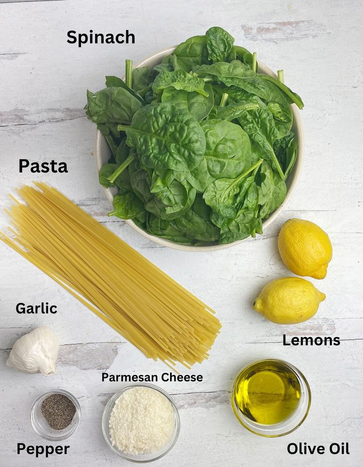 ingredients to make pasta laid out in bowls on a white wooden table with lemons, spinach, parmesan cheese, garlic and oil